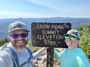 Logan S. posing at Crow Peak Summit sign with his wife.