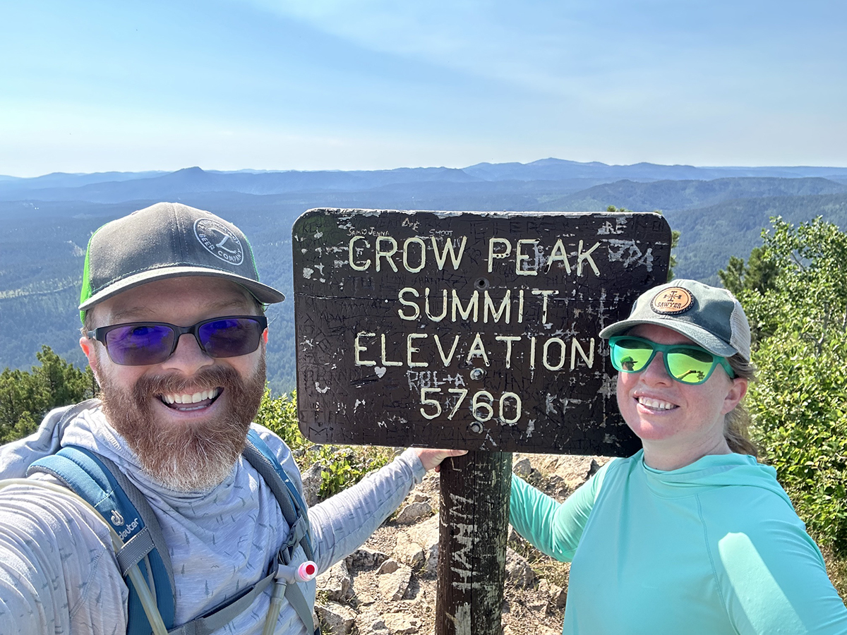 Logan S. posing at Crow Peak Summit sign with his wife.